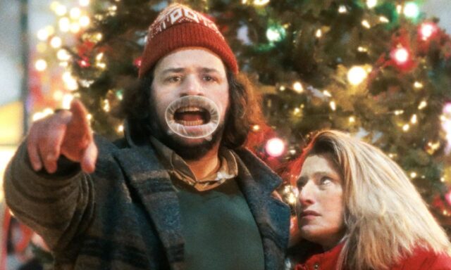 Man in silly hat in front of xmas tree mugging for camera while a woman looks on, rolling her eyes