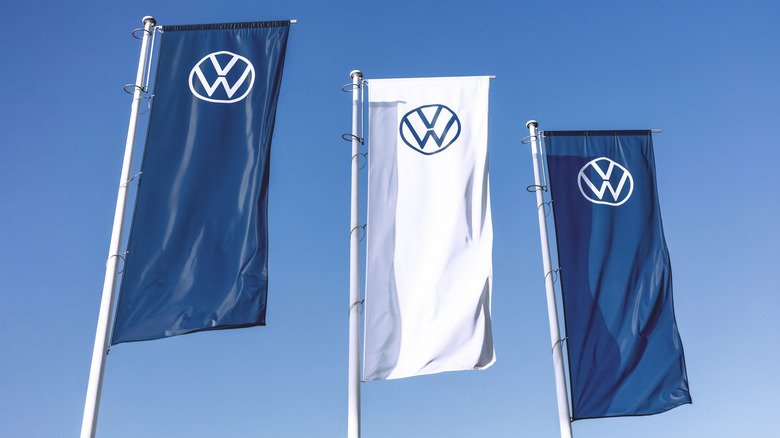 Photo of three tall flags displaying the Volkswagen logo in front of a daytime sky. The outer flags are blue with a white logo, while the middle flag is reversed.