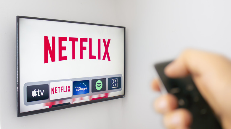 A blurred out hand holding a remote pointed towards a TV on a white wall showing Netflix and other streaming icons.