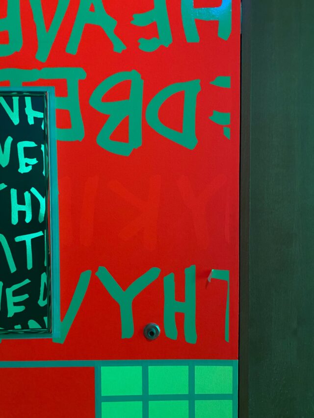 A close-up of a fluorescent mural at Newfields showing bright red DayGlo paint in an area formerly protected from light by the green vinyl lettering.
