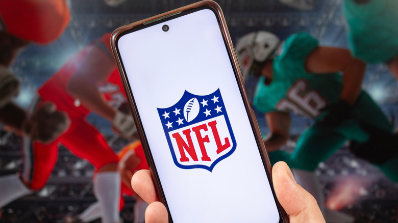 A man holding a smartphone with an NFL logo on the screen against the blurred backdrop of players rushing the football.