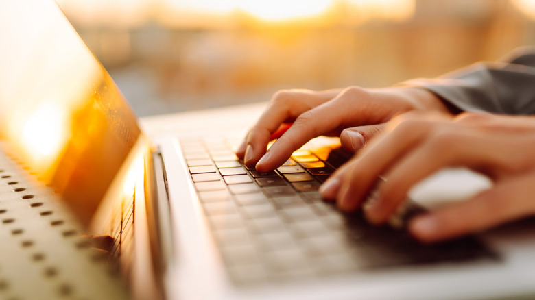Close-up of a person typing on a laptop outdoors in bright sunlight, with strong glare reflecting off the screen.